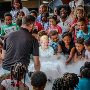 A group of children smile and lean in to observe a science demonstration producing clouds of vapor, led by "Mr. Science." 