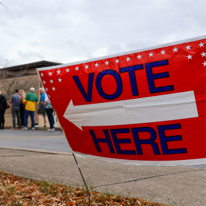 Vote Here sign in the foreground. People stand in line waiting to vote in the background.
