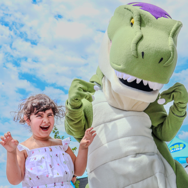 Young girl stands next to large costumed character at the summer kick off parties. Start planning your Summer at the Library.