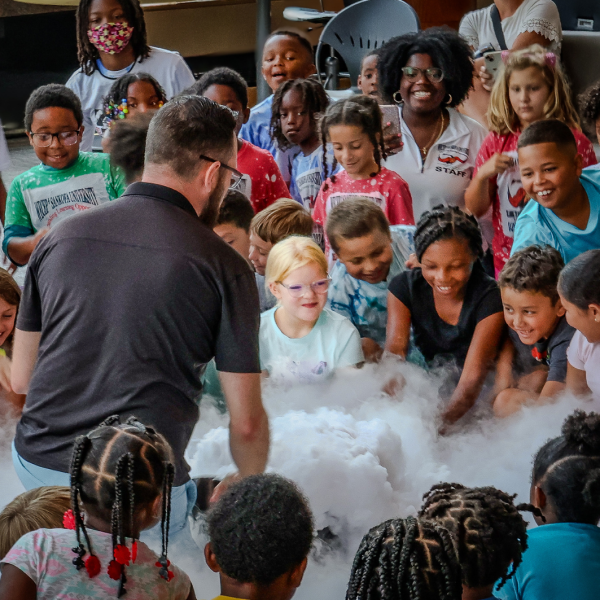 A group of children smile and lean in to observe a science demonstration producing clouds of vapor, led by "Mr. Science."