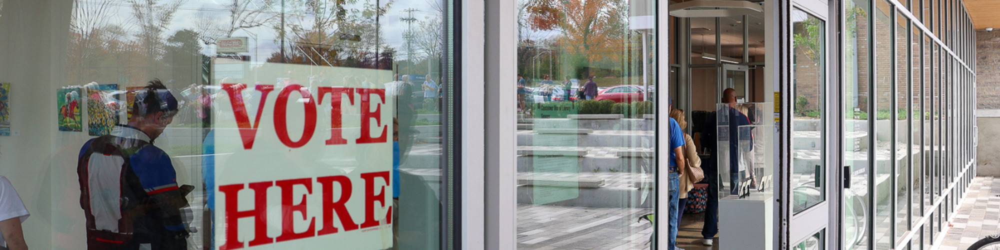 Photo of a sign that says "Vote Here" hanging in the window of a library branch, with people standing in line inside the building to vote
