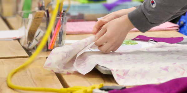 Hands hold fabric, an iron is featured in the right hand corner, and a yellow extension chord lays on a workspace table for crafting