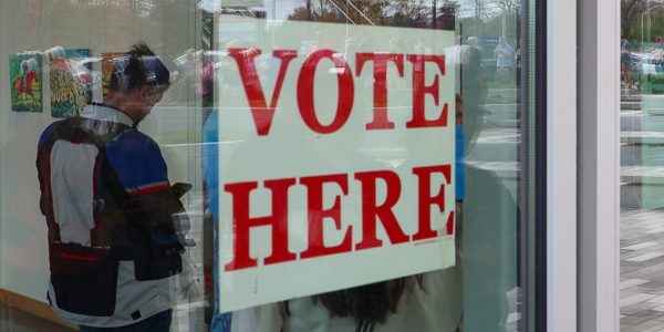 "Vote here" sign hangs in the window at the Marksbury Family Branch. A line of people getting ready to vote are in the background. 